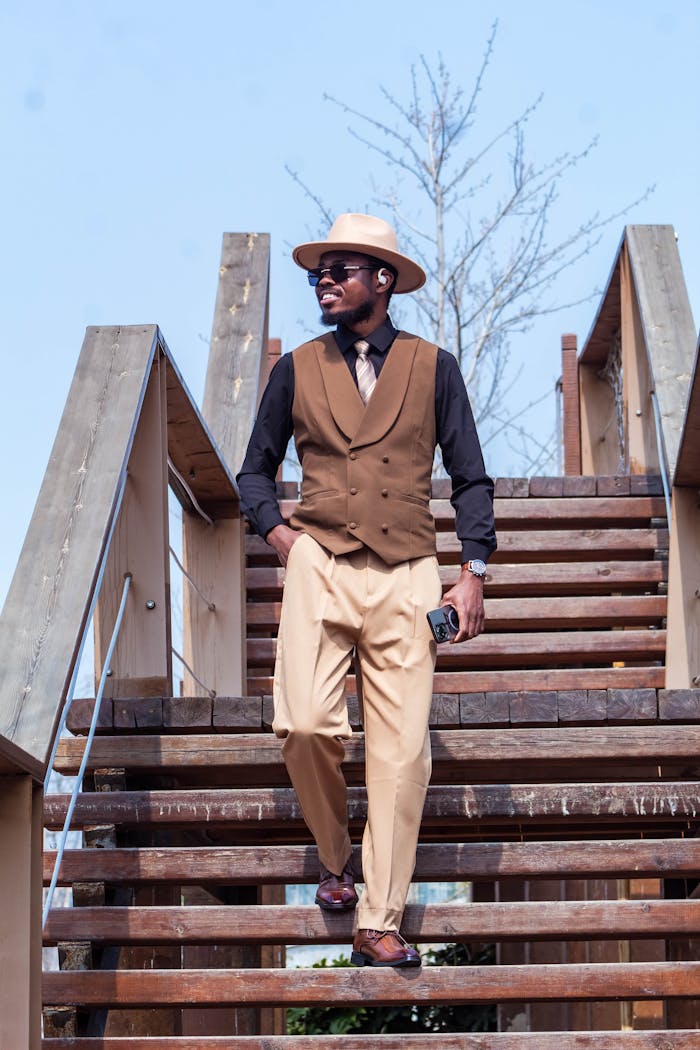 Sharp-dressed man in a vest and hat descends wooden stairs outdoors, embodying modern fashion.